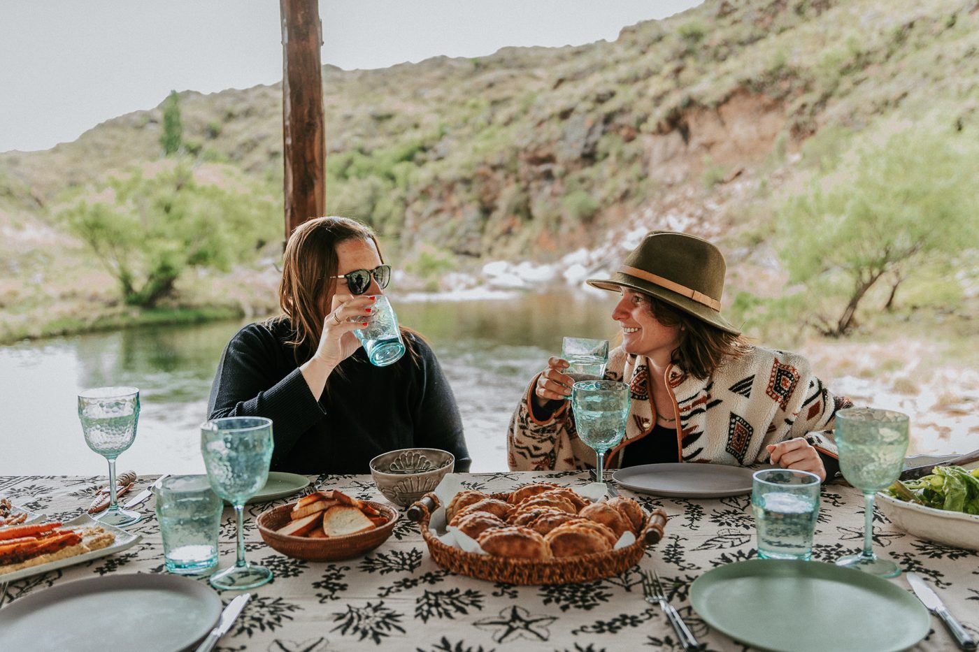 Two friends raise a glass at the quincho table by the river