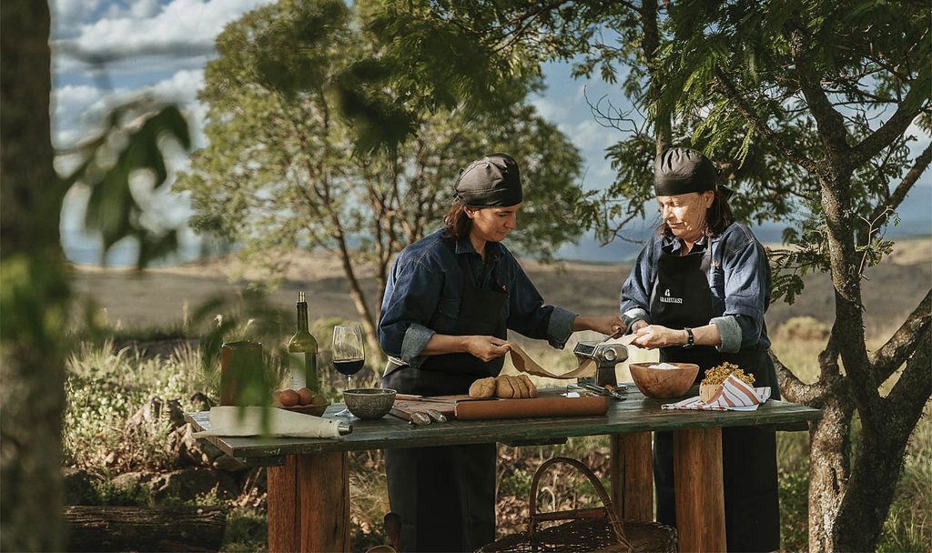 Two cooks preparing empanadas at an outdoor table, mountains and native trees in the background